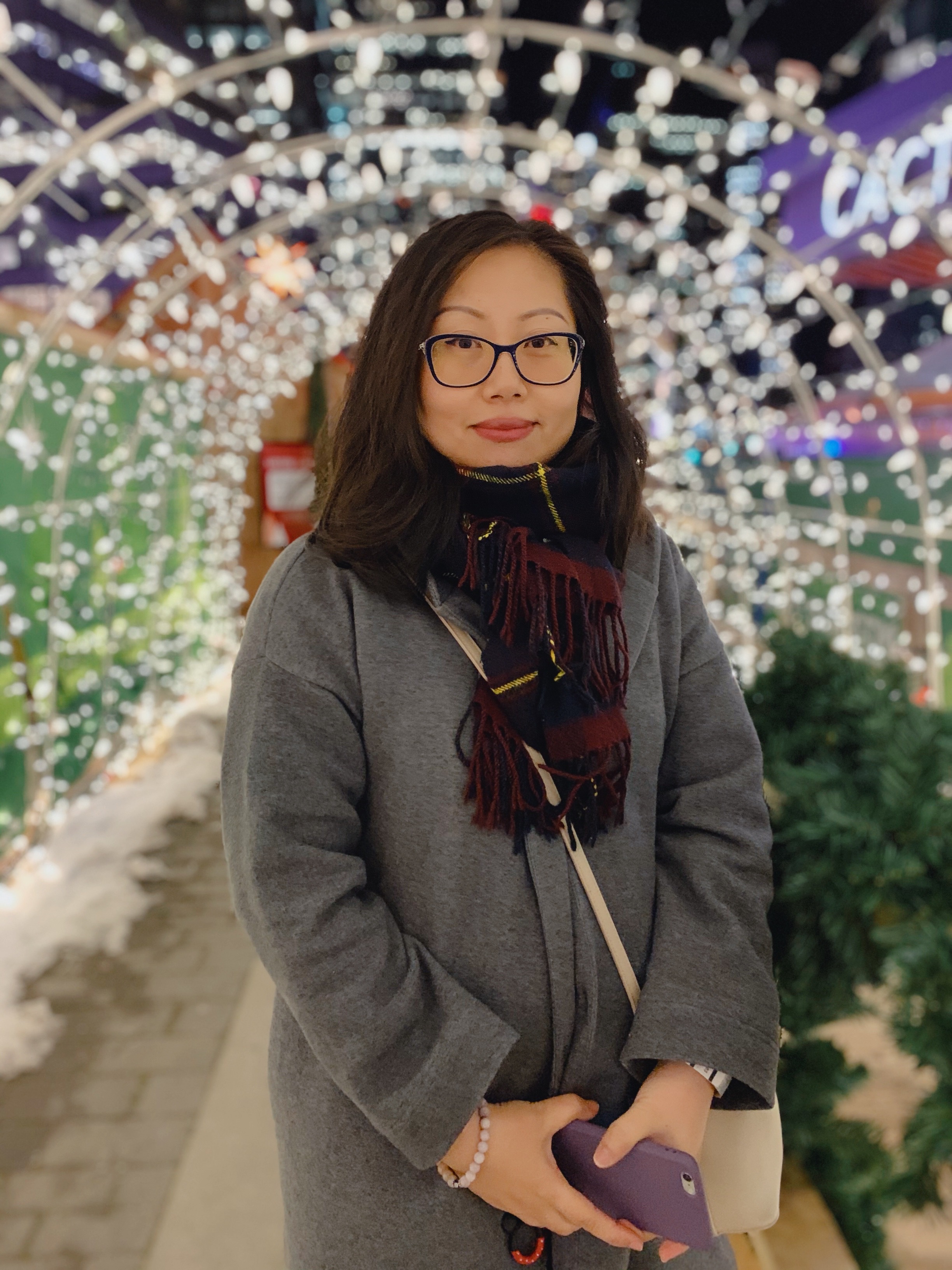Mingji is standing in the middle of the photo wearing a scarf and a winter's jacket. There are lights around her and a pathway behind her.